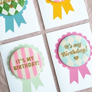 May include: Four white cards with colorful paper circles attached. Each circle has a different pattern and says "It's my Birthday!" in gold glitter letters. The circles are attached to a ribbon.