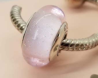 May include: A pink glass bead with silver glitter and a silver metal charm. The bead is on a silver bracelet.