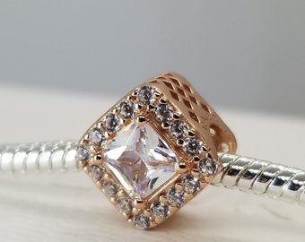 Puede incluir: Un charm de oro rosa con un cristal transparente cuadrado rodeado de cristales transparentes más pequeños.