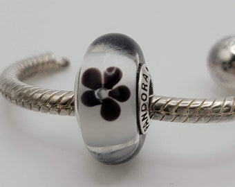 May include: A silver charm bracelet with a black and white glass bead featuring a flower design. The bead is inscribed with the word "PANDORA".