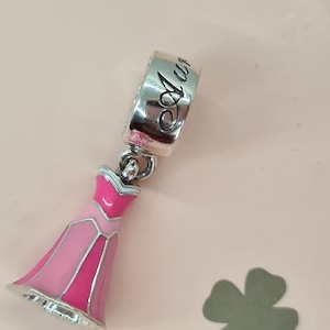May include: A silver charm with a pink and white enamel dress and the word "Aurora" engraved on the back.