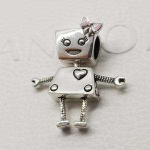 May include: A silver charm in the shape of a robot with a pink bow on its head. The robot has a heart on its chest and a happy face.