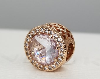 Puede incluir: Un charm de oro rosa con una piedra clara y facetada rodeada de un halo de piedras más pequeñas. El charm tiene un diseño delicado e intrincado.