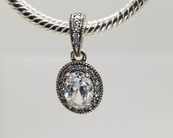 Puede incluir: Un charm de plata con un gran cristal transparente de forma ovalada rodeado de cristales transparentes más pequeños.