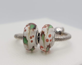 May include: Two glass beads with a white background and red and green holly leaf designs. The beads are on a silver bracelet.