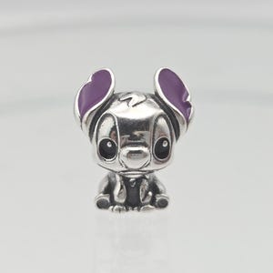 May include: A silver-colored charm featuring a cartoon character with large, purple-tipped ears and black eyes. The charm is a detailed, three-dimensional representation of the character, with a small, dark-colored nose and a slight smile. The charm is likely made of metal.