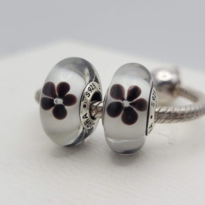 May include: Two silver charm beads with clear glass and a black flower design. The beads are on a silver bracelet.