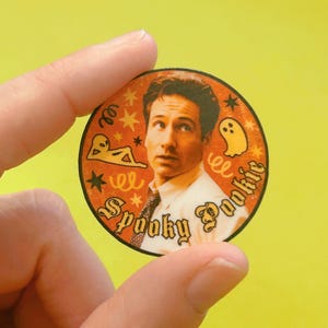 May include: A round, orange sticker with the text "Spooky Pookie" in a gothic font. The sticker features an image of a man with a surprised expression, along with illustrations of a ghost, an alien, and stars.