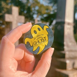 May include: A circular sticker with a yellow ghost design and the words "I'd rather be watching GHOST ADVENTURES" on a dark blue backdrop with white stars. The sticker is held up in front of a blurred outdoor background.