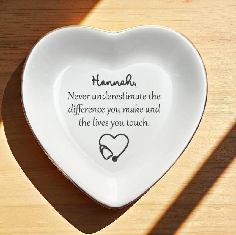 May include: A heart-shaped white ceramic dish with a gold rim. The dish features the name "Hannah" and the message "Never underestimate the difference you make and the lives you touch." A heart with a stethoscope is also displayed.