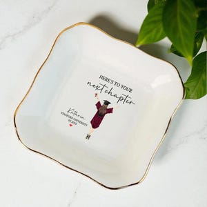 May include: A white ceramic dish with a wavy edge and gold trim. The dish features the text "HERE'S TO YOUR next chapter" and a graduation illustration. The name "Kathrin" and "Stanford University 05.2025" are also printed on the dish.