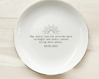 Może przedstawiać: Biały ceramiczny talerz ze złotą obwódką. Talerz posiada czarny wzór promieni słonecznych i tekst: "May every sunrise provide more strength and every sunset bring more peace. 09/02/2025."