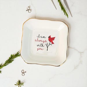 Può includere: Un vassoio quadrato in ceramica bianca con bordo dorato. Il vassoio presenta il testo "I am always with you" e un'illustrazione di un uccello cardinale rosso. Elementi decorativi sparsi intorno al vassoio.