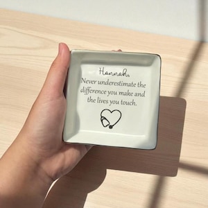 May include: A square, off-white ceramic trinket dish with a black rim. The dish features the text "Hannah, Never underestimate the difference you make and the lives you touch." A heart and stethoscope graphic are below the text.