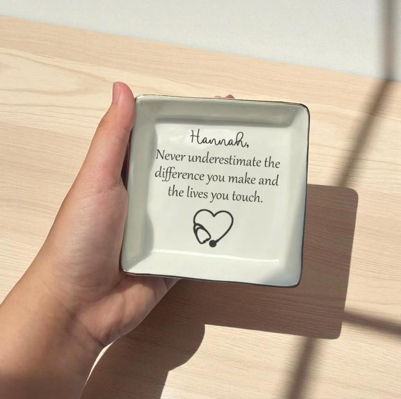 May include: A square, off-white ceramic trinket dish with a black rim. The dish features the text "Hannah, Never underestimate the difference you make and the lives you touch." A heart and stethoscope graphic are below the text.