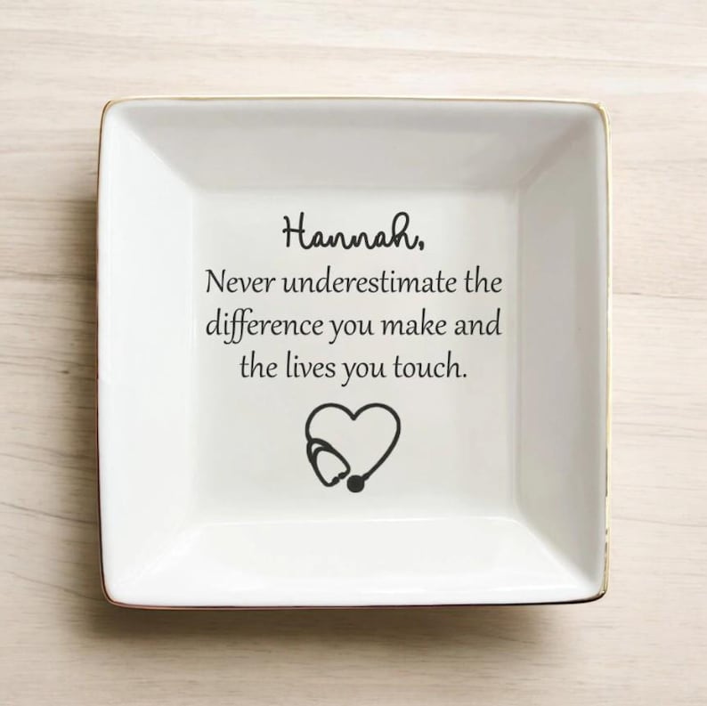 May include: A white square ceramic dish with a gold rim. The dish has the name "Hannah" written in cursive, along with the message "Never underestimate the difference you make and the lives you touch." A heart-shaped stethoscope graphic is below the text.