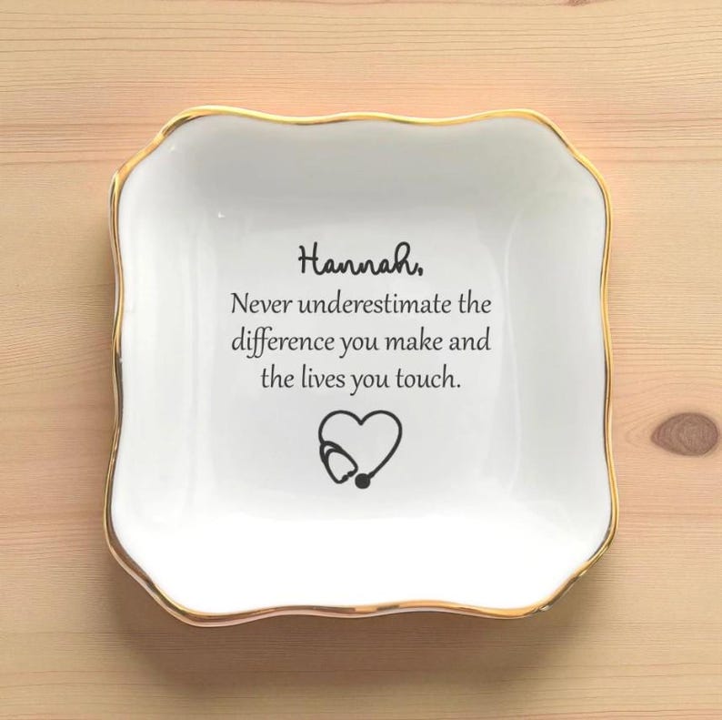 May include: A white ceramic trinket dish with a gold rim. The dish has a square shape with wavy edges. Text on the dish reads "Hannah, Never underestimate the difference you make and the lives you touch." A heart with a stethoscope is below the text.