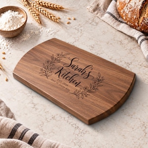 Op de afbeelding: Een rechthoekige donkerbruine houten snijplank met afgeronde randen, voorzien van een lasergegraveerd bloemenkransontwerp rondom de tekst Sarah's Kitchen, EST. 2026, en de zin love in every bite in het midden van het oppervlak.