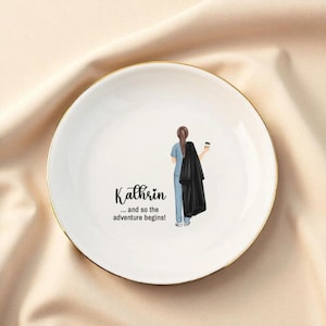 May include: White ceramic trinket dish with a gold rim. The dish features a graphic of a person in blue scrubs holding a black coat and coffee cup. The name "Kathrin" and the phrase "...and so the adventure begins!" are printed on the dish.