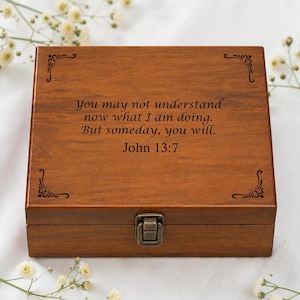 Könnte beinhalten: Eine rechteckige Holzschatulle mit dunkelbrauner Oberfläche und einem Metallverschluss an der Vorderseite. Der Deckel ist mit dem eingravierten Text You may not understand now what I am doing. But someday, you will. John 13:7 und Verzierungen versehen.