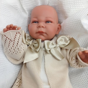 May include: A baby doll wearing a cream knitted cardigan with a satin bow. The doll has realistic features and blue eyes.