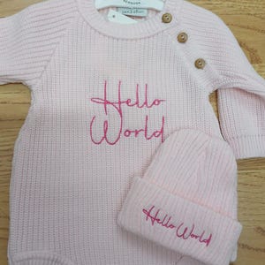 May include: A pale pink knitted baby romper and beanie set. The romper has long sleeves, wooden buttons, and the embroidered text "Hello World" in pink. The beanie also features the embroidered text "Hello World".