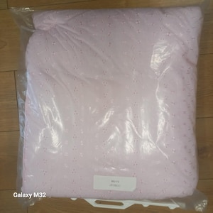 May include: A pale pink, eyelet-patterned blanket is packaged in a clear plastic bag. The blanket features a delicate, textured surface with small, evenly spaced holes. A small white label is visible on the bag.