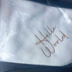 May include: White baby blanket with the embroidered text "Hello World" in a brown script font. The blanket is in a clear plastic bag.