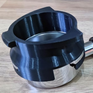 Puede incluir: Un portafiltro negro para máquina de espresso con una cesta de filtro de acero inoxidable y un mango negro brillante. El portafiltro está sobre una superficie de madera, con una pegatina "Made in the UK".