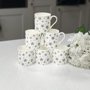 May include: Six white ceramic mugs with a grey star pattern. The mugs are stacked in a pyramid shape on a white countertop.