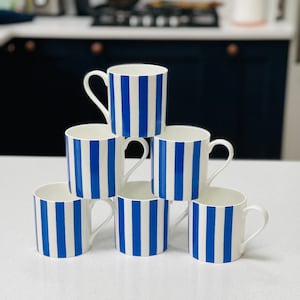 May include: Six white ceramic mugs with blue stripes stacked in a pyramid shape on a white countertop.