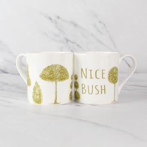 Może przedstawiać: Dwa białe ceramiczne kubki z zielonym wzorem drzewa. Na jednym kubku widnieje napis "NICE BUSH" w kolorze zielonym. Drugi kubek ma wzór drzewa. Kubki mają zakrzywiony uchwyt.