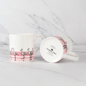 Op de afbeelding: Twee witte keramische mokken met flamingo-ontwerpen. Elke mok heeft een roze gestreepte band met flamingo-illustraties. Eén mok staat rechtop, de andere is gekanteld en onthult de tekst "Finsbury China" op de bodem.