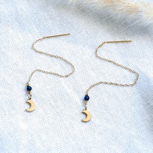 Könnte beinhalten: Ein Paar goldfarbene Ohrhänger. Jeder Ohrring hat eine feine Kette, eine kleine dunkelblaue Perle und einen Mondsichel-Anhänger. Präsentiert auf einer weißen, strukturierten Oberfläche.