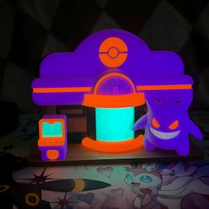 May include: A glowing purple and orange light-up display featuring a building with a circular emblem at the top, a bright turquoise central entrance, and a small arcade machine. A purple Gengar figure with glowing orange eyes stands to the right.