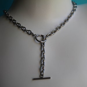 May include: A silver chain necklace with a T-bar closure. The chain is a curb style and has a round ring at the center.