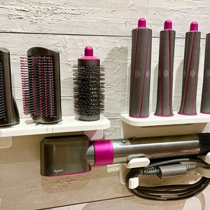 May include: A white wall-mounted organizer with a Dyson hair dryer, a round brush attachment, and four curling iron attachments. The organizer has a white shelf and a gray cord holder.