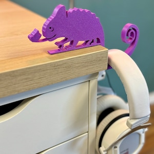 May include: Purple 3D printed chameleon headphone holder on a wooden desk with a white drawer. The chameleon has a long tail that wraps around the headphones.