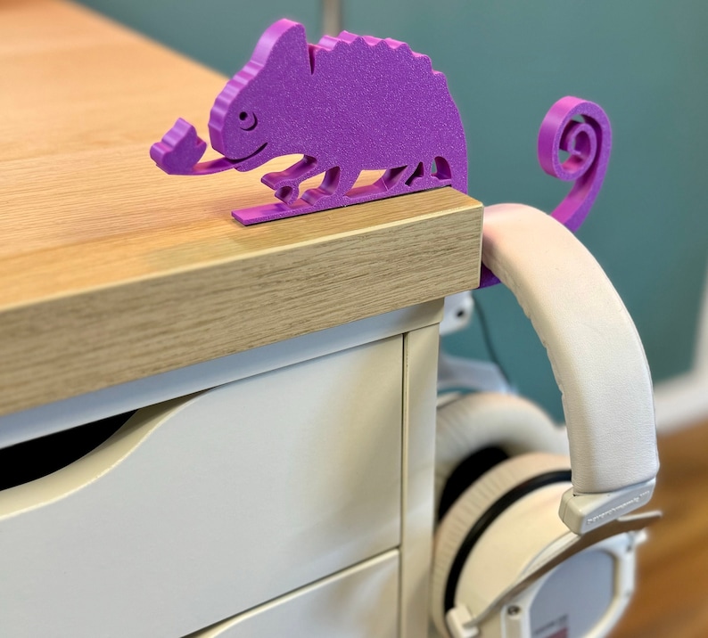 May include: Purple 3D printed chameleon headphone holder on a wooden desk with a white drawer. The chameleon has a long tail that wraps around the headphones.