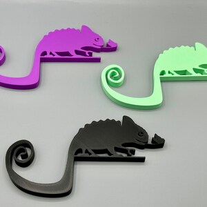 May include: Three chameleon-shaped phone holders in purple, green, and black. Each holder has a curled tail and a small, raised detail on the chameleon's head.