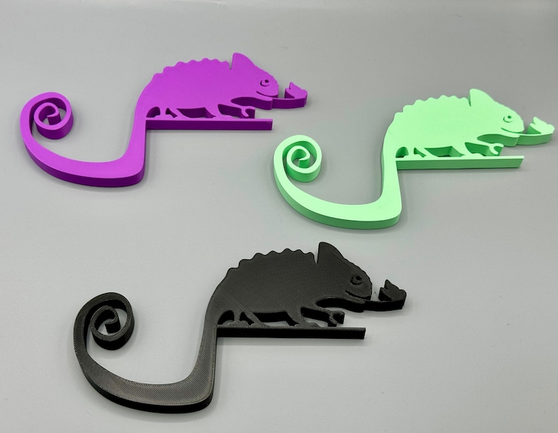 May include: Three chameleon-shaped phone holders in purple, green, and black. Each holder has a curled tail and a small, raised detail on the chameleon's head.