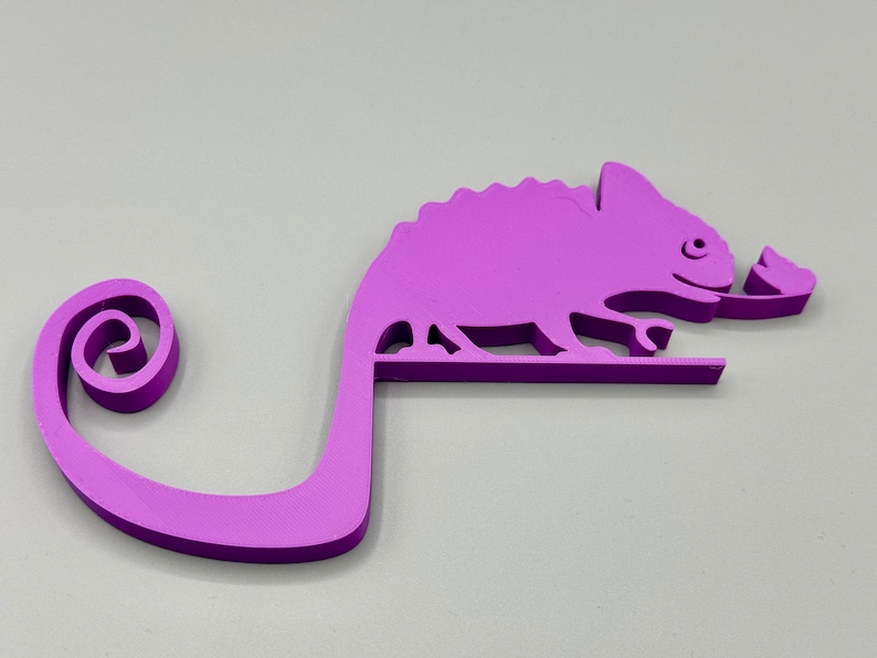 May include: A purple 3D printed chameleon figurine with a curled tail. The chameleon is holding a leaf in its mouth.