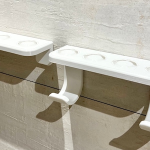 May include: Two white plastic toothbrush holders mounted on a wall. The holder on the left has three toothbrush slots, while the holder on the right has four toothbrush slots. Both holders have a hook on the bottom.