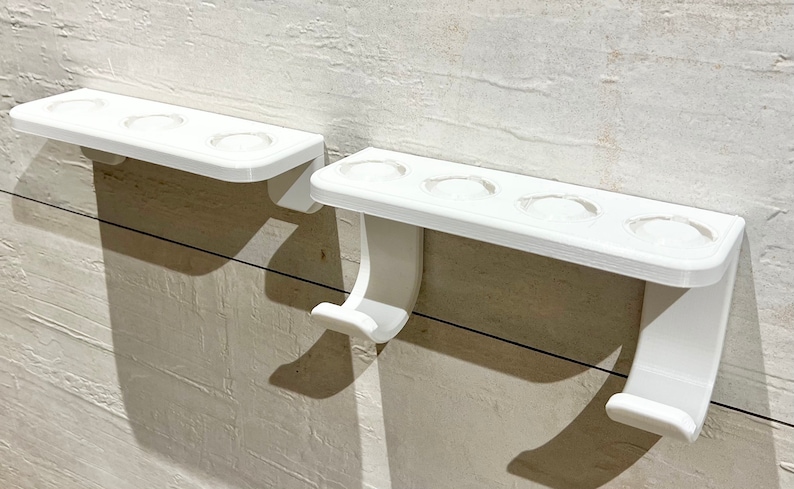 May include: Two white plastic toothbrush holders mounted on a wall. The holder on the left has three toothbrush slots, while the holder on the right has four toothbrush slots. Both holders have a hook on the bottom.