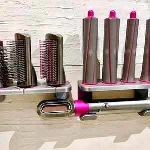 May include: A set of hair styling tools with a pink and silver design. The set includes a hair dryer, a round brush, two styling brushes, and four curling barrels. The tools are mounted on a wall-mounted stand.