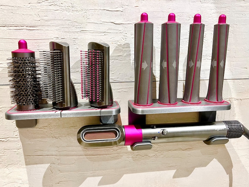 May include: A set of hair styling tools with a pink and silver design. The set includes a hair dryer, a round brush, two styling brushes, and four curling barrels. The tools are mounted on a wall-mounted stand.