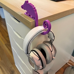 May include: Purple 3D printed chameleon headphone holder with a white pair of headphones hanging from it.