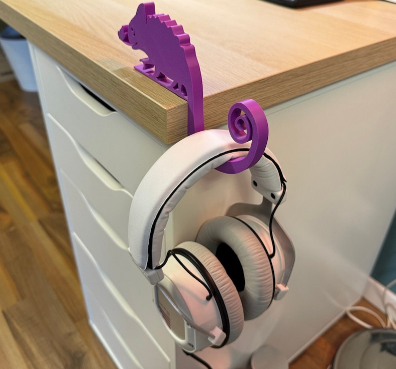 May include: Purple 3D printed chameleon headphone holder with a white pair of headphones hanging from it.