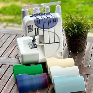 May include: A white overlock sewing machine with a blue and grey control panel. The machine is on a wooden table with five colourful plastic bobbin cases in front of it.