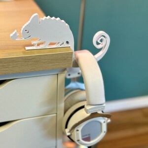 May include: White 3D printed chameleon headphone stand with a curled tail. The stand is mounted on a white desk with drawers. A pair of white headphones are hanging from the stand.